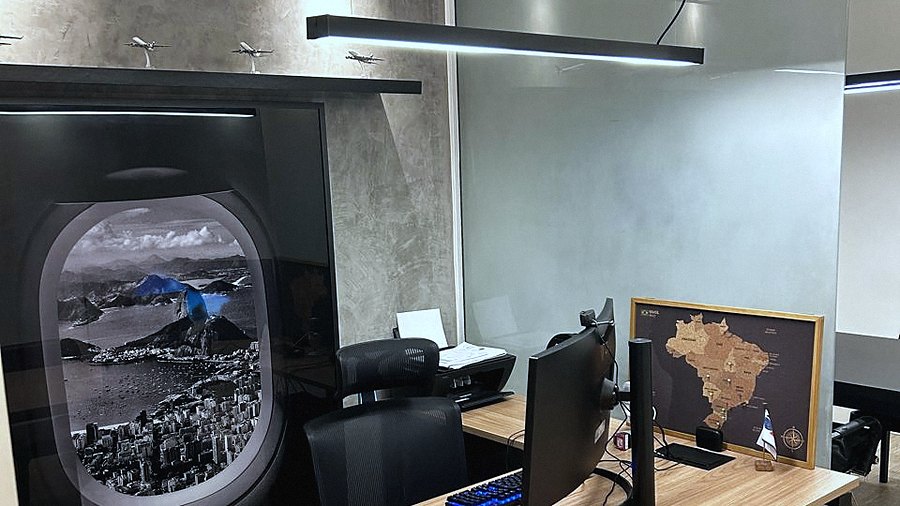 Estação de trabalho do escritório: painel em forma de janela de avião com a vista do Rio, miniaturas de aeronaves na prateleira e um mapa do Brasil em cortiça sobre a mesa.