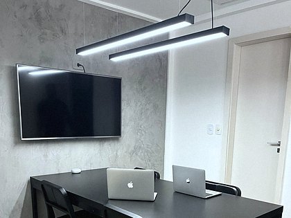 Sala de reunião com mesa preta, televisor na parede de concreto e luminárias lineares.